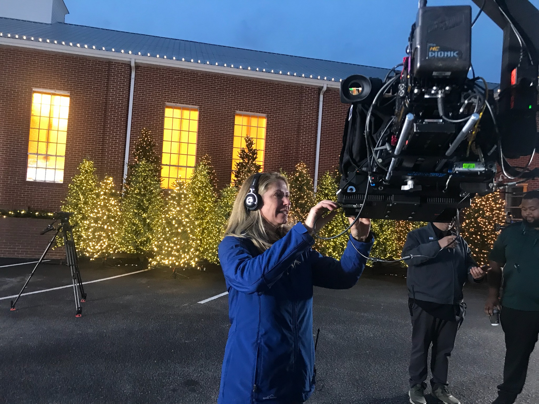 B Prompt operator adjusting a jib-mounted camera and teleprompter at dusk outside a holiday-decorated venue