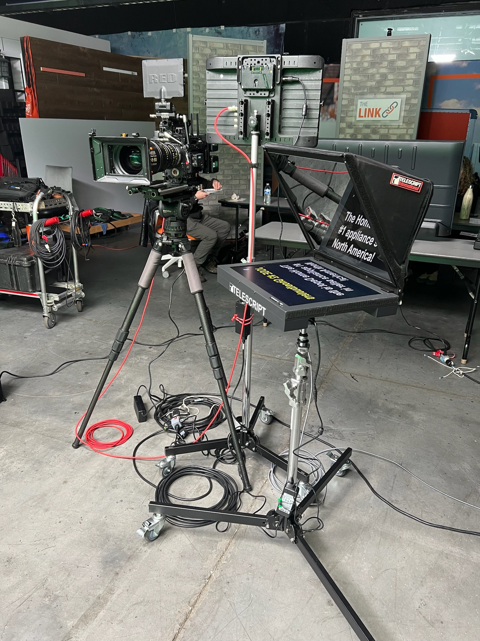 RED cinema camera on a tripod beside a Telescript International teleprompter on a rolling stand in a studio set