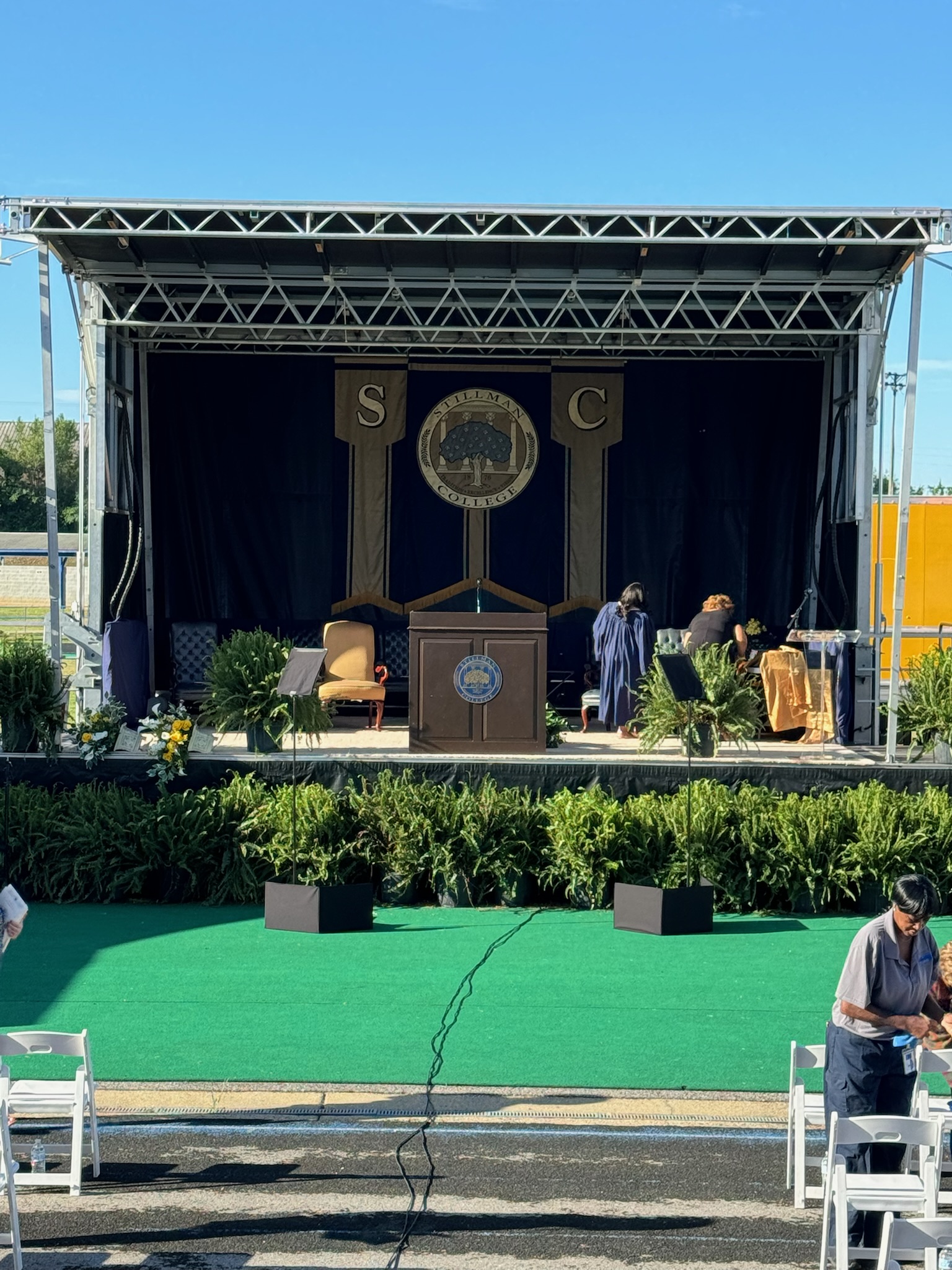Outdoor commencement stage with Stillman College banners, podium, and ferns under a blue sky